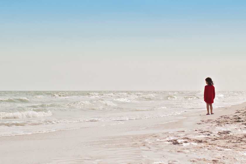 A photo showing a little girl in red standing alone on a beach.